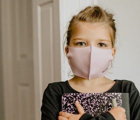 Niña pequeña con una mascarilla puesta en la cara y sujetando un cuaderno